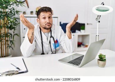 Young Doctor Working At The Clinic Using Computer Laptop Clueless And Confused Expression With Arms And Hands Raised. Doubt Concept. 