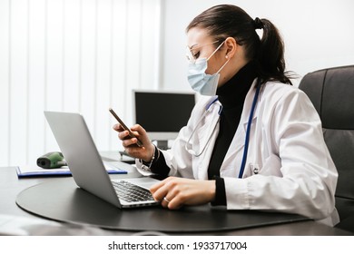 Young Doctor Working On Laptop In Office Analyzes Medical Records Of His Patients Infected Whit Coronavirus Covid-19 Wearing Protective Face Mask Take A Break Looking A Message On Smartphone