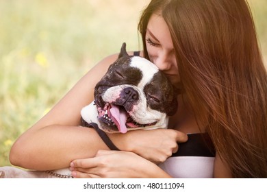 Young Dog Owner Girl Plays With French Bulldog Puppy In Park Outdoor.Teen Brunette Female Kissing Little Puppy.Domestic Canine Breed.Young Woman Cuddling Her Pet Doggy