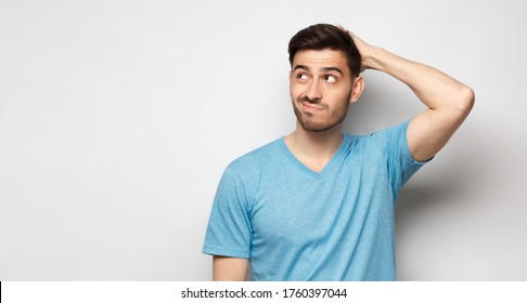 Young doubtful man thinking, scratching head and trying to find solution, isolated on gray background