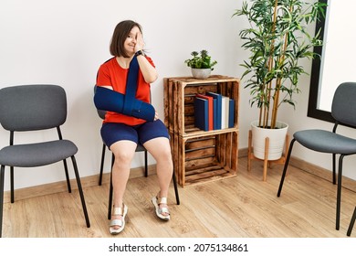 Young Down Syndrome Woman Sitting At Doctor Waiting Room With Arm Injury Covering One Eye With Hand, Confident Smile On Face And Surprise Emotion. 