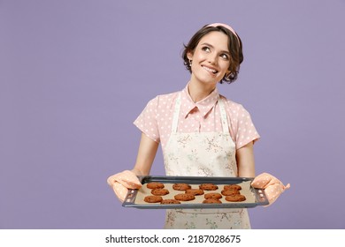 Young Dreamful Housewife Housekeeper Chef Cook Baker Woman Wearing Pink Apron Showing Chocolate Cookies Biscuits On Baking Sheet Look Aside Isolated On Pastel Violet Background. Cooking Food Concept.