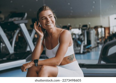 Young dreamful minded happy strong sporty athletic sportswoman woman 20s in white sportswear earphones listen music warm up training sit near treadmill look aside in gym indoors Workout sport concept. - Powered by Shutterstock