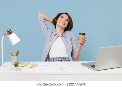 Young dreamful secretary employee business woman in casual shirt sit work at white office desk with pc laptop hold cup of coffee hold hand behind neck head isolated on pastel blue background studio.