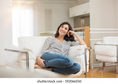 Young dreaming woman sitting on sofa at home. Thoughtful young woman sitting in living room and looking up. Smiling woman relaxing on couch in a modern home. - Powered by Shutterstock