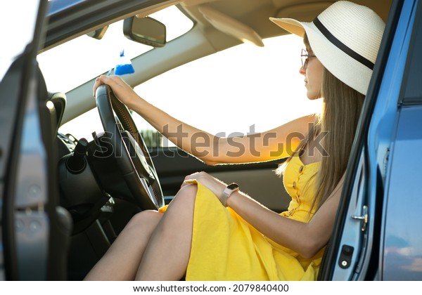 Young driver girl wearing yellow dress and straw\
hat sitting behind steering wheel driving a car. Summer vacation\
and travel concept.