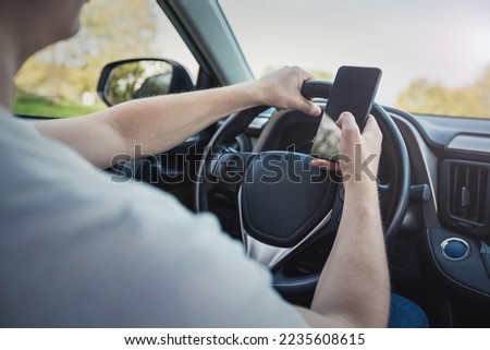 Young driver guy distracted by his phone while in front of the steering wheel, using his smartphone with one hand while driving. Risk and danger situations on the road, violating traffic rules
