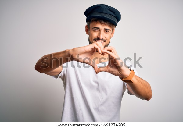 Young driver man with beard\
wearing hat standing over isolated white background smiling in love\
showing heart symbol and shape with hands. Romantic\
concept.