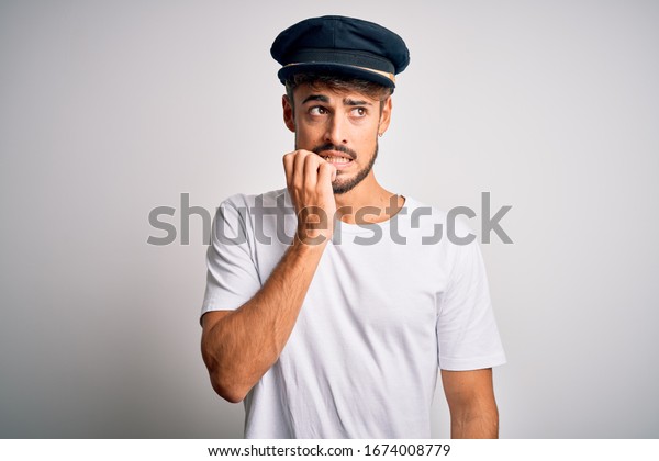 Young driver man with beard\
wearing hat standing over isolated white background looking\
stressed and nervous with hands on mouth biting nails. Anxiety\
problem.