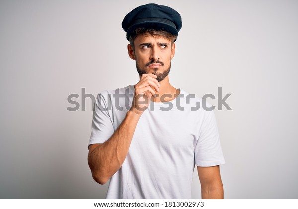 Young driver man with beard wearing hat standing\
over isolated white background with hand on chin thinking about\
question, pensive expression. Smiling with thoughtful face. Doubt\
concept.