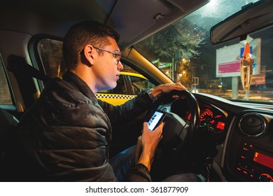 Young Driver Using His Smartphone While Driving. Irresponsible Teenager Texting And Driving At Night In The City.