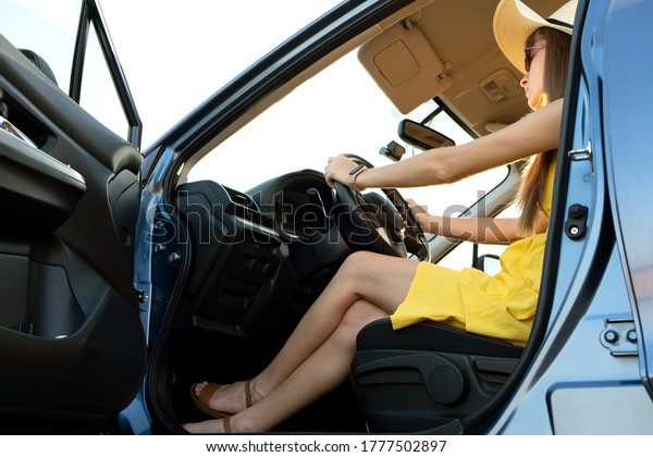 Young driver woman wearing yellow dress and\
straw hat sitting behind steering wheel driving a car. Summer\
vacation and travel\
concept.