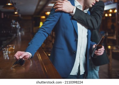 Young drunk businessman is holding a bottle of beer and reaching car keys on bar counter in pub, another man is stopping him