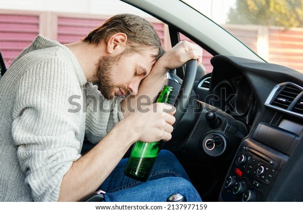 Young drunk man fall asleep\
behind the wheel of a car. Male car driver holding bottle of\
beer.