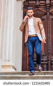 Young East Indian American Businessman With Beard, Wearing Brown Blazer, White Round Collar Shirt, Blue Pants, Holding Laptop Computer, Walking Down Stairs From Office Door Way, Talking On Cell Phone.