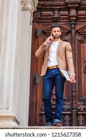 Young East Indian American Businessnan With Beard, Wearing Brown Blazer, White Round Collar Shirt, Blue Pants, Holding Laptop Computer, Standing In Front Old Style Office Door, Talking On Cell Phone.