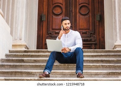 Young East Indian American College Student With Beard Studying In New York, Wearing White Shirt, Blue Pants, Sitting On Stairs Outside On Campus, Working On Laptop Computer, Talking On Cell Phone.