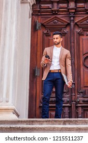 Young East Indian American Man With Beard, Wearing Light Brown Blazer, White Round Collar Shirt, Blue Pants, Holding Laptop Computer, Standing In Front Old Style Door Way, Texting On Cell Phone.