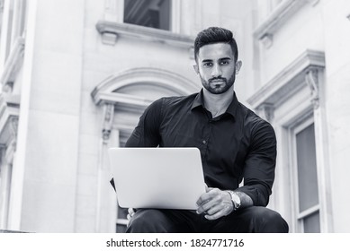 Young East Indian American Man With Beard, Studying, Working In New York City, Wearing Black Shirt, Holding Laptop Computer, Sitting Outside Old Style Office Building, Looking Up. Black And White.
