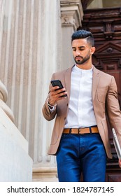 Young East Indian American Man With Beard, Wearing Brown Blazer, White Round Collar Shirt, Blue Pants, Carrying Laptop Computer, Standing By Old Style Doorway, Looking Down, Texting On Cell Phone.
