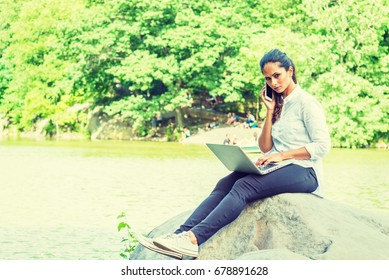 Young East Indian American Woman Wearing White Shirt, Black Pants, White Sneakers, Sitting On Rocks By Lake At Central Park, New York, Working On Laptop Computer, Looking Up, Talking On Cell Phone.
