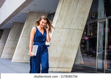 Young Eastern European American Woman talking on cell phone, traveling, working in New York City, wearing blue sleeveless jumpsuit, carrying laptop computer, walking on street outside office building. - Shutterstock ID 1178585734