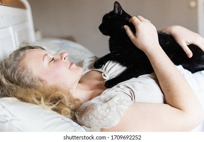 Young Eastern European Woman Lying In Bed With Black Cat In Her Hands In The Morning.