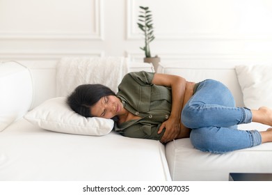 A Young Eastern Woman Lying On The Couch And Suffers From Month Menstruation Period Pain, Stomach Cramps, Indian Young Female Is Holding Tummy With A Grimace Of Misery On Her Face