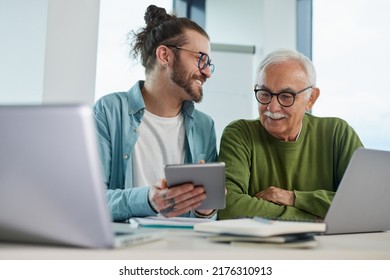 Young Educator Showing Senior Student How To Solve A Problem On Project On His Tablet While The Student Typing On A Laptop.