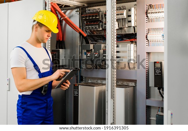 A young electrician works with a\
tablet standing near an industrial fuse board\
cabinet.