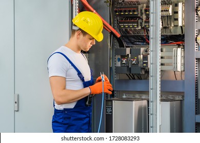A young electrician in a yellow hard hat cuts the blue wire with pliers. An employee maintains a switchboard at the factory. Indoor. - Shutterstock ID 1728083293