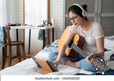 Young Elegant Asian Woman In Headphones With Tablet Computer Sitting On Bed Play Acoustic Guitar At Home. Girl Musician Love Music Making Writing New Songs On Weekends Holidays Stay At Cozy Room