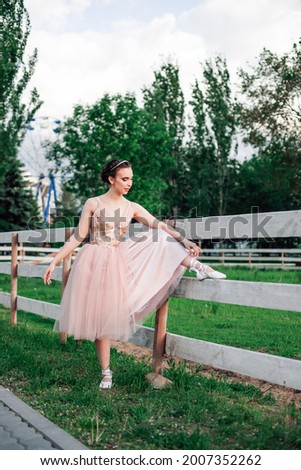 a young, elegant ballerina has lifted her foot on the wooden board of the fence of the horse pen in the amusement park and is adjusting the satin ribbons on her ballet flats.