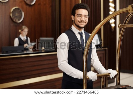 Young elegant bellboy in uniform pushing cart with clients baggage while moving along hotel lounge and looking at camera