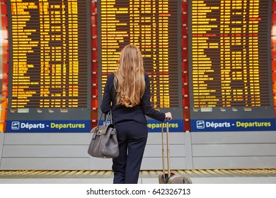 Young elegant business woman with hand luggage in international airport terminal, looking at information board, checking her flight. Cabin crew member with suitcase.