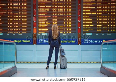 Young elegant business woman with hand luggage in international airport terminal, looking at information board, checking her flight. Cabin crew member with suitcase.