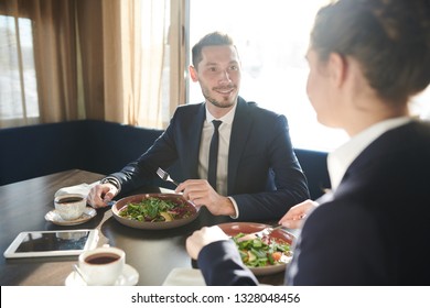 Young elegant businessman eating fresh vegetable salad at lunch break with his colleague or partner in front