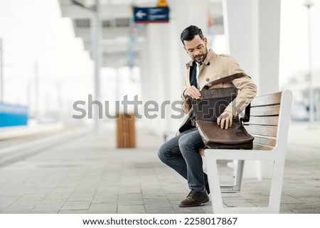 A young elegant businessman is sitting on a train station, waiting for a metro and searching for documents in a bag.