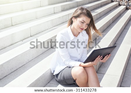 Young elegant businesswoman sitting outdoor, working on tablet. Sunny day.