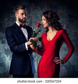 Young Elegant Couple In Evening Dress Portrait. Woman In Red Sniff Rose. Focus On Woman.