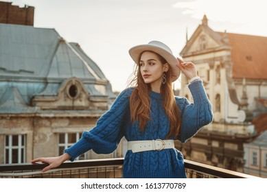 Young elegant fashionable  woman, model wearing trendy blue knitted sweater, wide leather belt, stylish white hat, earrings, posing at sunset, in European city. Copy, empty space for text
