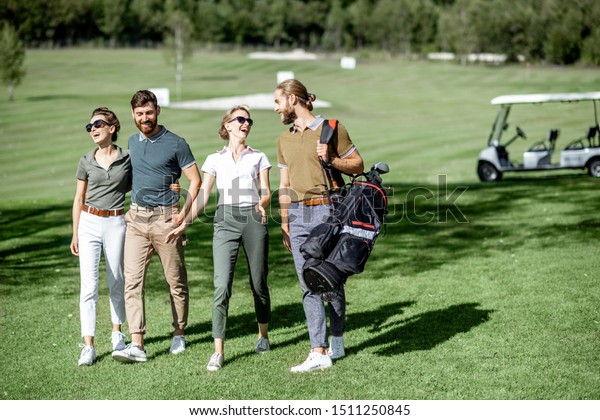 Young and elegant friends walking with golf\
equipment, hanging out together on the beautiful course with golf\
car on the background
