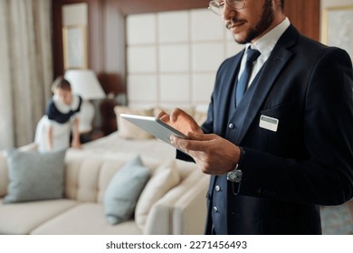 Young elegant manager of luxurious hotel using tablet against chambermaid changing bedclothes and preparing room for new guests