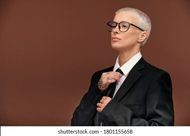 Young Elegant Masculine Female With Short Dyed Blond Hair Tying Black Necktie While Getting Dressed For Work Or Formal Event In Isolation