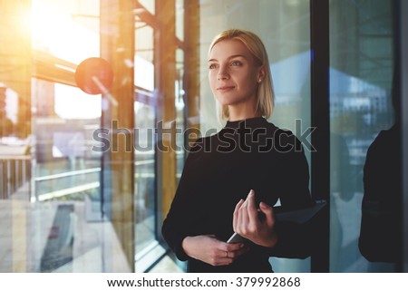 Young elegant woman thinking something while standing with touch pad near office window outside, female employer holding digital tablet while looking forward to meeting with international partners