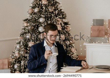 Young, elegantly dressed man sitting at a table, reading a book or journal and drinking coffee, with a dazzling, warmly lit Christmas tree and wrapped gifts in the background