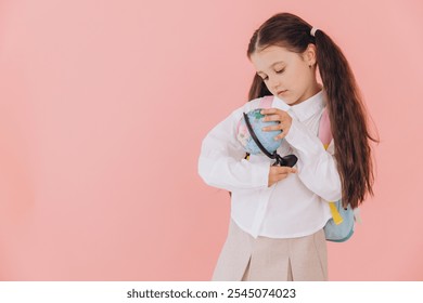 Young elementary school student in uniform with backpack, holding a small globe on a pink background, studying geography - Powered by Shutterstock