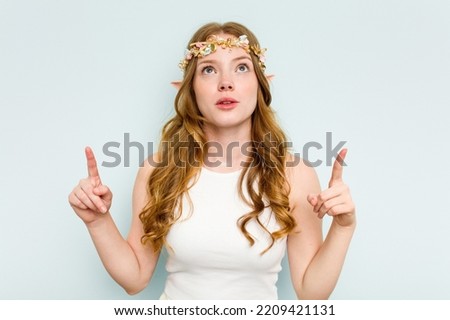 Young elf woman isolated on blue background pointing upside with opened mouth.