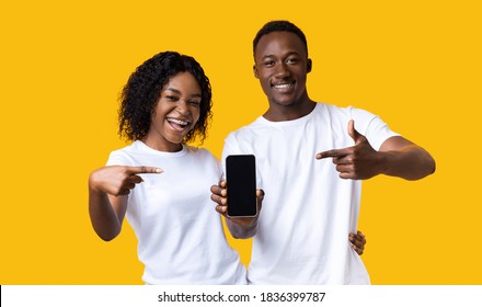Young Emotional African-american Couple Pointing Fingers At Blank Black Smartphone Screen, Yellow Studio Background. Smiling Black Man And Woman Showing Nice Application On Their Mobile Phone, Mockup