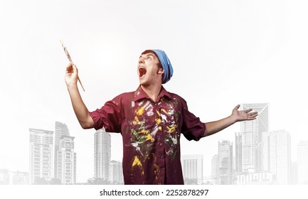 Young emotional artist with open mouth gesturing with paintbrush. Happy painter in shirt and bandana standing on background modern office buildings. Creative hobby and artistic occupation concept - Shutterstock ID 2252878297